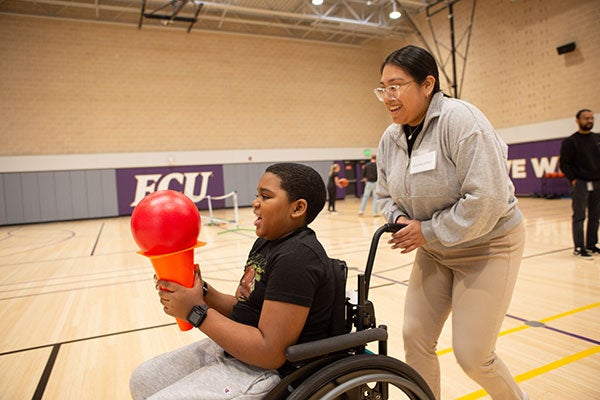 A boy in a wheelchair holds a ball while being pushed by a woman.