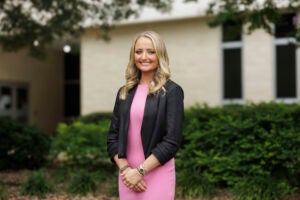 Hailey Lewis, wearing a pink dress and black jacket, stands in front of the Brody School of Medicine building.