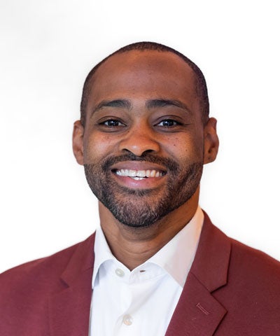 Professional headshot of a man with short hair and a trimmed beard, wearing a maroon jacket and white shirt, smiling at the camera against a plain white background.