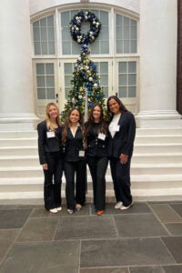 Four women in business attire smile in front of a decorated Christmas tree and a large building with white columns.