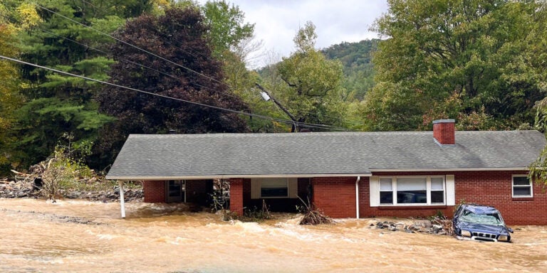 Fast-moving and turgid flood waters move down a moderate slope upon which a red brick ranch house sits partially submerged.