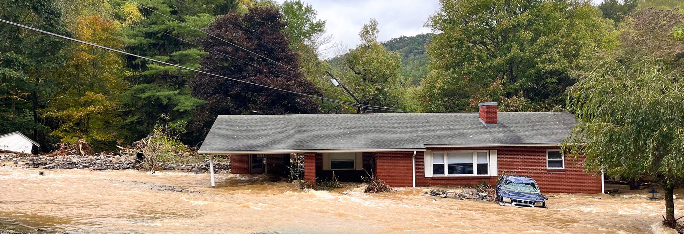 Fast-moving and turgid flood waters move down a moderate slope upon which a red brick ranch house sits partially submerged.