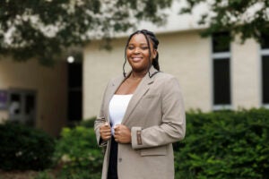 Kamryn Henderson, wearing a tan jacket and white blouse, poses with her hands on the lapels of her jacket.