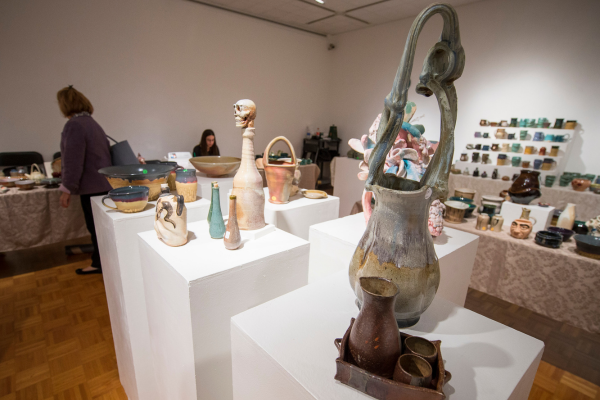 This image shows the interior of an art sale taking place in an art gallery. The space features various ceramic and pottery artworks displayed on white pedestals. The artworks include abstract sculptures, vases and other ceramic forms in various styles and colors. There are also some framed artworks and shelves of smaller ceramic pieces visible in the background.