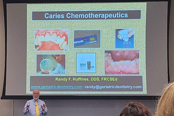 A speaker gestures as he talks in front of a slide depicting teeth as part of a presentation to a seated audience.