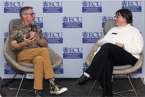 A man and woman are sitting across from one another and are holding microphones. They are sitting in front of a photo backdrop with the East Carolina University Thomas Harriot College of Arts and Sciences logo.