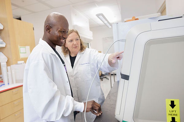 Paul Lovens and Elizabeth Ables, both in white lab coats, stand next to a monitor in a science lab on campus.