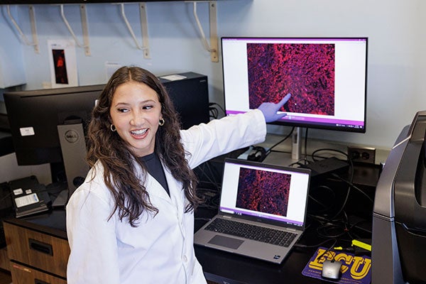 Bayli Locklear, in a white lab coat, stands in front of a laptop and monitor, pointing at red and purple cells on the screen.