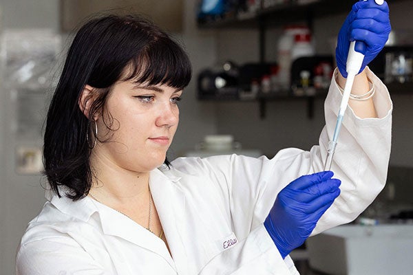 Ellisa Fisher, wearing a white lab coat and blue latex gloves, holds up a tool in the lab and adds a solution to a glass test tube.