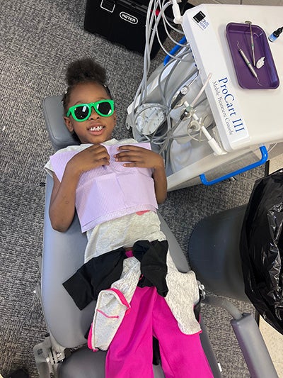 A young girl wearing protective glasses sits reclined in a dental operatory chair with her mouth open wide as a hygienist works on her teeth with instruments.