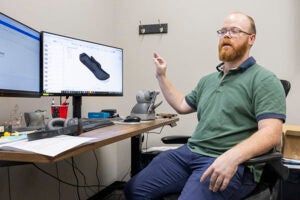A man in a green shirt gestures with his right hand as he sits near a computer screen that shows a protective pad that attaches to the bottom of a prosthetic foot.