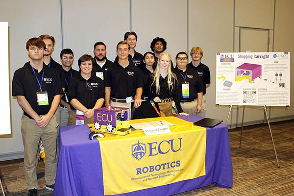 Twelve people in black shirts stand behind a table that includes a purple and gold robot that looks like a small, covered shopping cart. Next to them is a poster that describes how they built the robot. The table is covered with a purple and gold tablecloth.