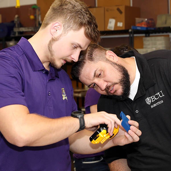 A male college student wearing a purple shirt at left holds a plastic part as another male college student wearing a black shirt leans in to examine the part. 