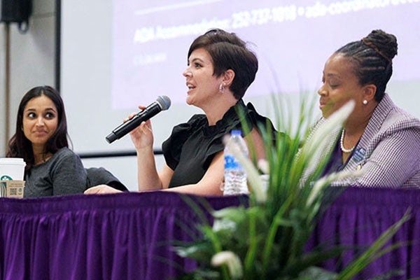 Jessica Doyle-Mekkes, wearing a black shirt, holds a microphone and talks while sitting at a table between two women.