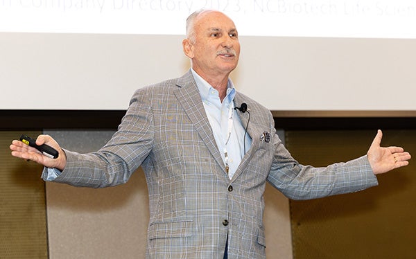 A man in a gray sport coat outstretches both of his arms as he speaks in front of a screen from a stage.