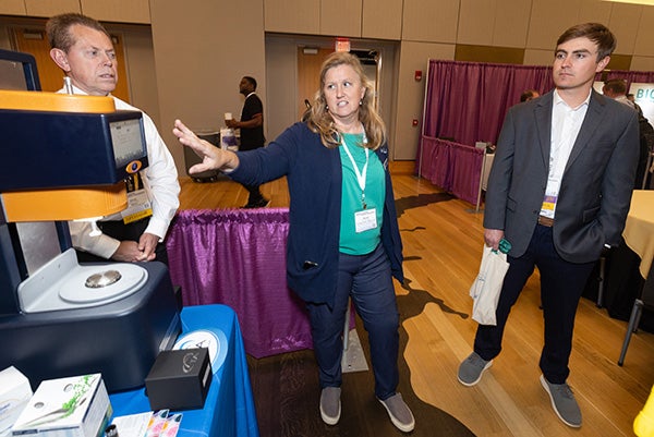 A woman at center gestures toward a piece of equipment as a man in a white shirt at left stands behind the equipment and a man at right in a sport coat listens.