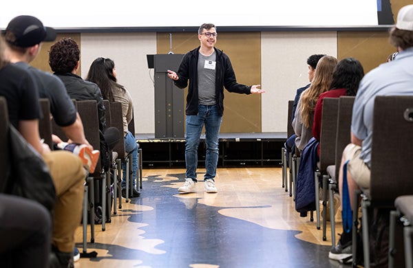 A man wearing a black jacket and gray shirt gestures with his hands as he stands and talks to numerous people seated in chairs to the left and right of him.