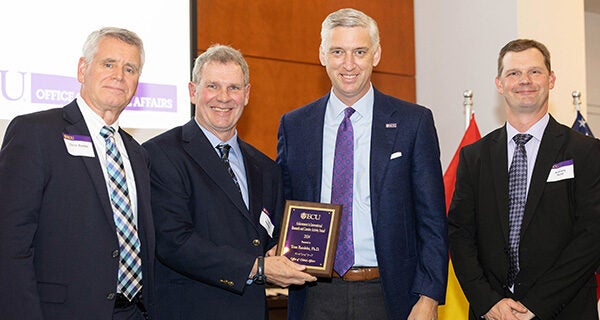 Four middle-aged men in suits pose for the camera. One holds a plaque. 