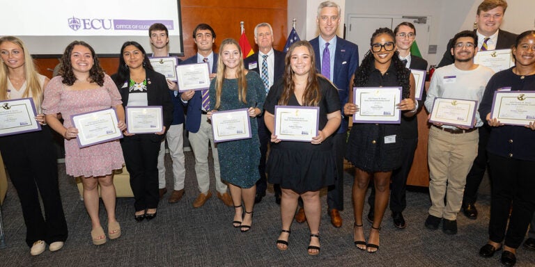 A diverse group of 15 students in business casual attire pose for a photo holding certificates with three men in suits.