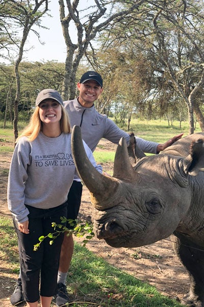 A woman and a man pose with a rhinoceros in a wooded area in Kenya.