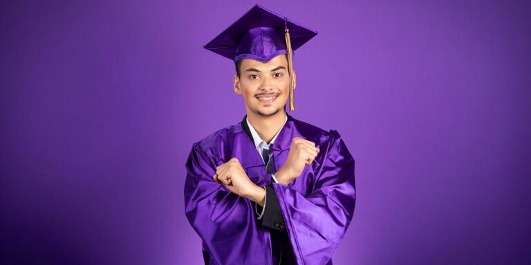 A young man with a mustache and goatee wears a purple mortarboard with gold tassel and purple graduation gown crosses his arms with closed fists while smiling.