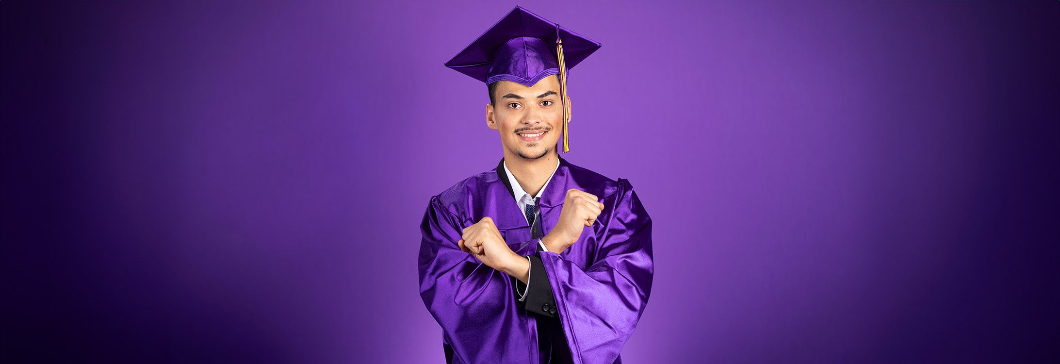 A young man with a mustache and goatee wears a purple mortarboard with gold tassel and purple graduation gown crosses his arms with closed fists while smiling.