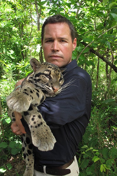 A man with short brown hair, wearing a blue button-down shirt and khaki pants, holds a baby leopard and is standing in front of a forest of green foliage.