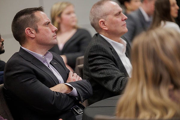  a man in a suit jacket and collared shirt watches on with crossed arms as students present their business ideas.