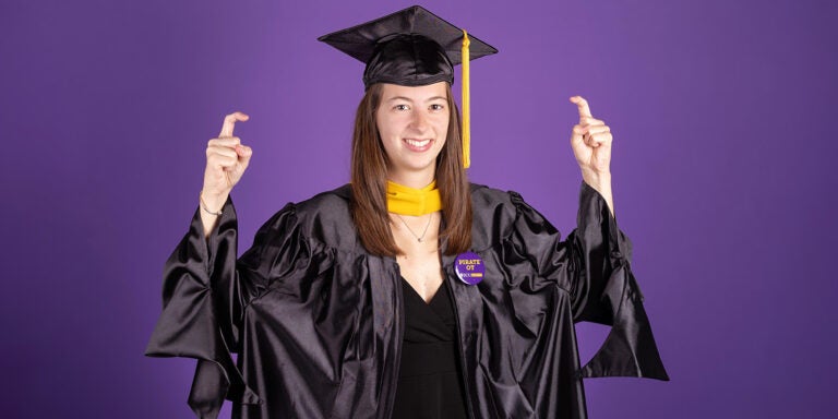 A woman wearing a graduation cap and gown holds her fingers in the shape of a hook.