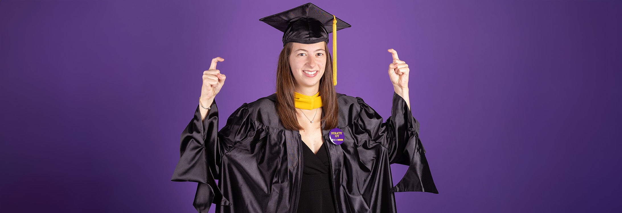 A woman wearing a graduation cap and gown holds her fingers in the shape of a hook.