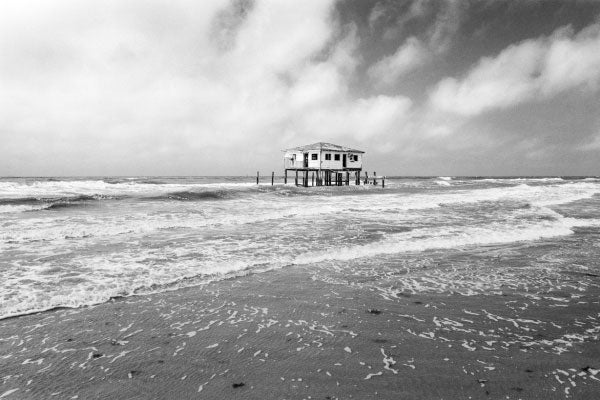 In black and white, a small building on stilts surrounded by water, with beach sand in the foreground and right corner.