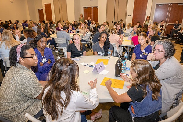 Men and women sit around round tables in a conference room setting talking with one another and taking notes on colored sheets of paper.