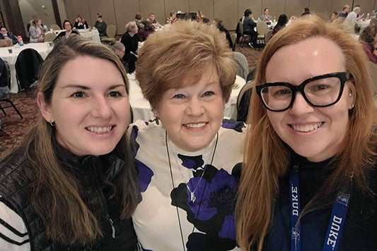 Three women pose for a photo in a ballroom filled with attendees seated at conference tables.