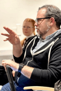 A man with facial hair and glasses sits and speaks, holding what looks like a tablet in his left hand, and gesturing with his right.
