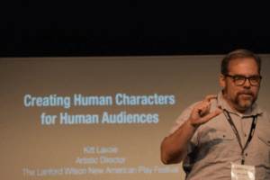 A man wearing a lanyard nametag speaks in front of a projection screen showing the text, “Creating Human Characters for Human Audiences, Kitt Lavoie, Artistic Director, The Lanford Wilson New American Play Festival.”