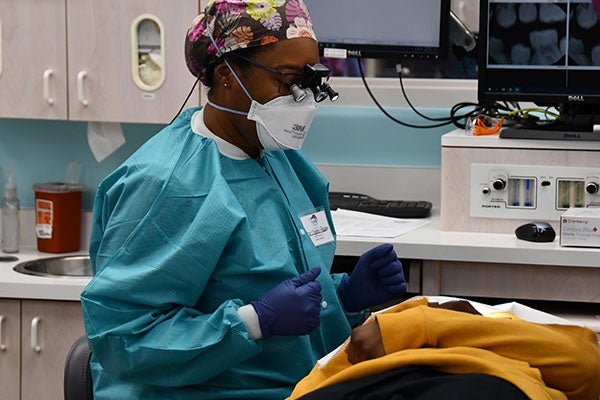 A dentist holds dental tools and sits over a pediatric patient reclined in a treatment chair awaiting care.