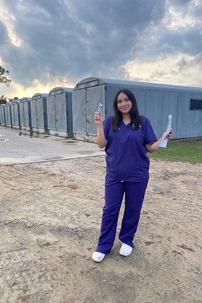 A woman with dark hair wearing purple medical scrubs holds medicine at a farm setting with metal buildings in the background.