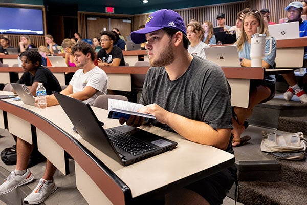 : Students sit at desks, and in front of computers, in a large stadium seating classroom. A male student closest to the camera in a purple ECU hat is reading a book while listening to the instructor.