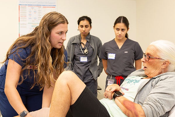 A woman with long brown hair wearing blue medical scrubs speaks with an elderly woman in a medical clinical setting as two other women stand in the background. The elderly woman is wearing makeup that simulates bruising on her head and arms.