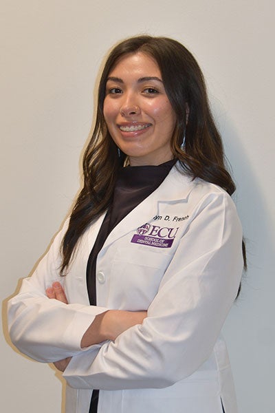 A dental student stands with arms crossed, wearing her white coat embossed with her name.