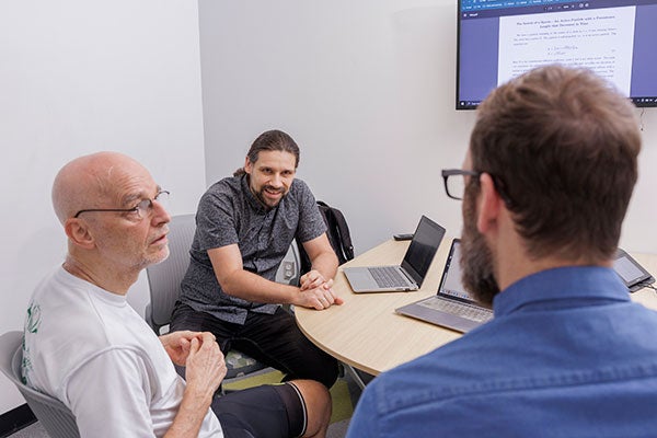 A man in a grey shirt smiles while talking to a man in a white shirt. A man wearing a blue shirt has his back to the camera.