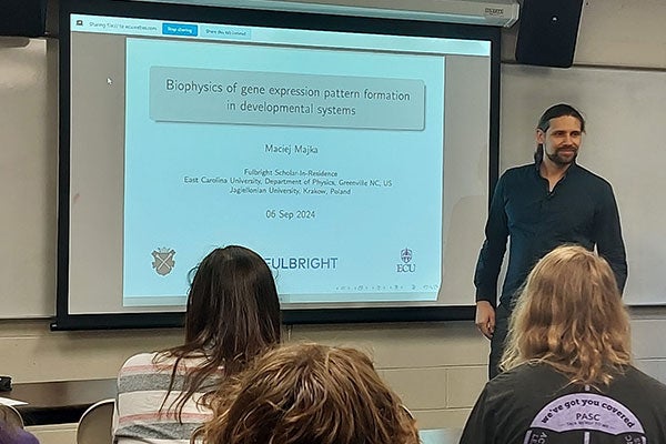 A man in a blue shirt stands in front of a projector screen in a classroom as people look on.