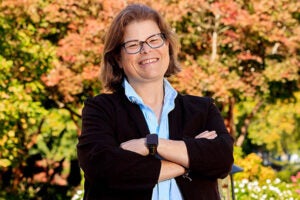 A woman with short brown hair and glasses wearing a black blazer and blue shirt stands with her arms crossed in front of a background of trees.