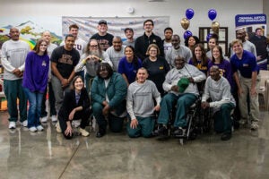A group of students kneel and stand inside a room with a concrete floor and purple and gold balloons and the Department of Criminal Justice and Criminology banner in the background.
