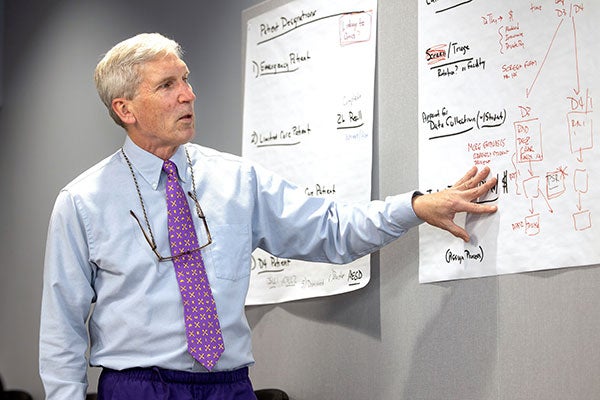 A man in a blue shirt and purple tie points to words on a piece of paper during a lecture.