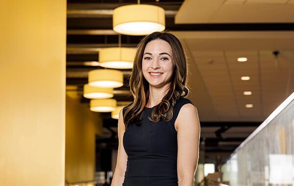 A young woman in a black dress stands in a yellow hallway smiling at the camera.