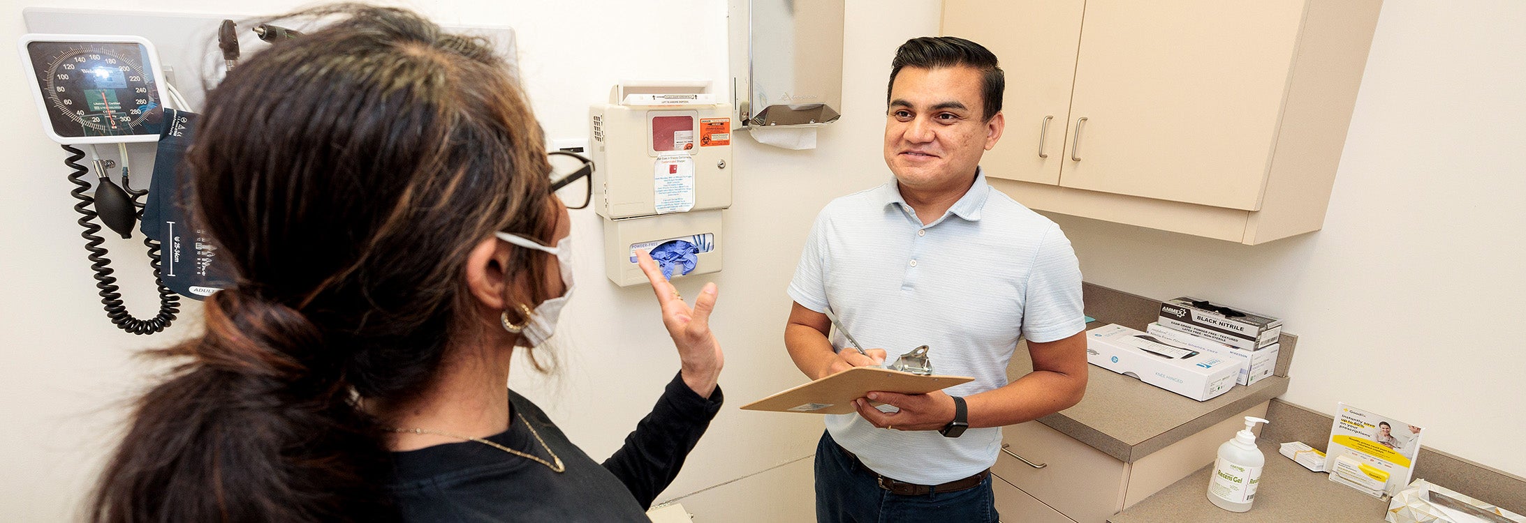 A male medical provider in a blue shirt holding a clipboard consults with a patient in an exam room