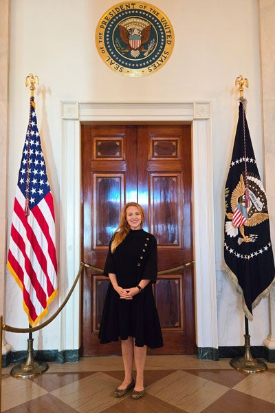 A woman in a dark dress with a row of gold buttons smiles as she stands in front of a large wooden door flanked by two flags and crowned with the American presidential seal.