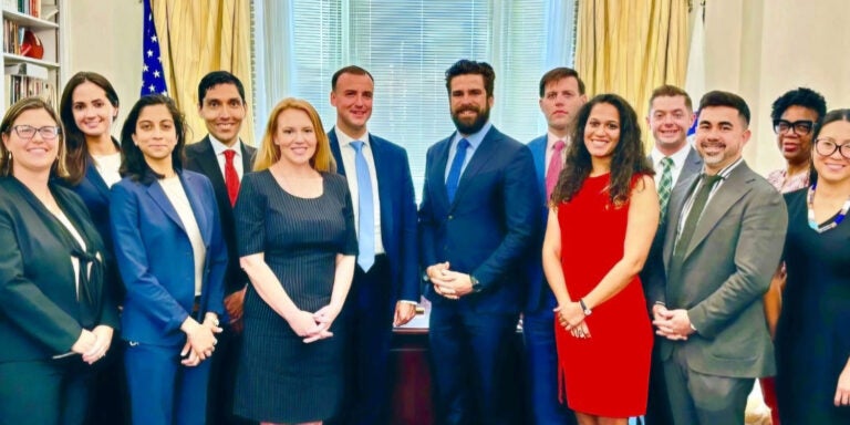 A group of 16 women and men in dress clothes pose in an office with a large wooden desk and gold drapes framing a window.