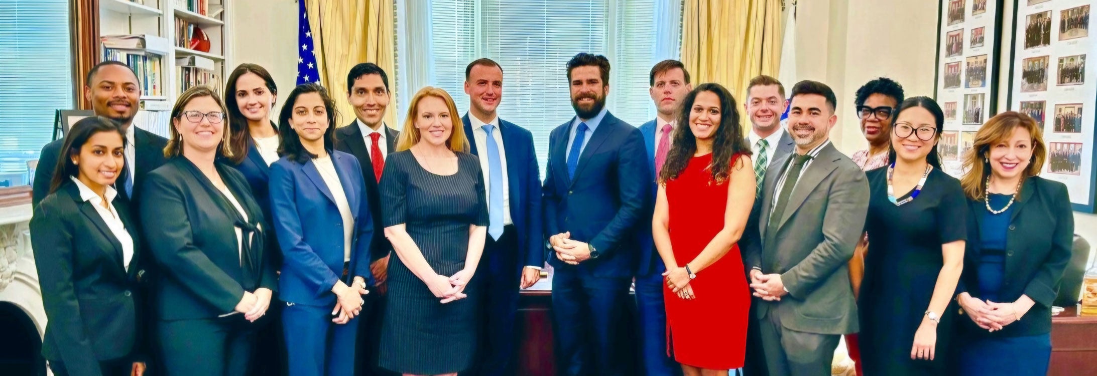 A group of 16 women and men in dress clothes pose in an office with a large wooden desk and gold drapes framing a window.